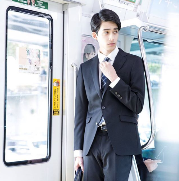 ブラックスーツを着用した男性が、電車の中のドア付近に立っている。ネクタイ着用。結び目のあたりを触っている。
